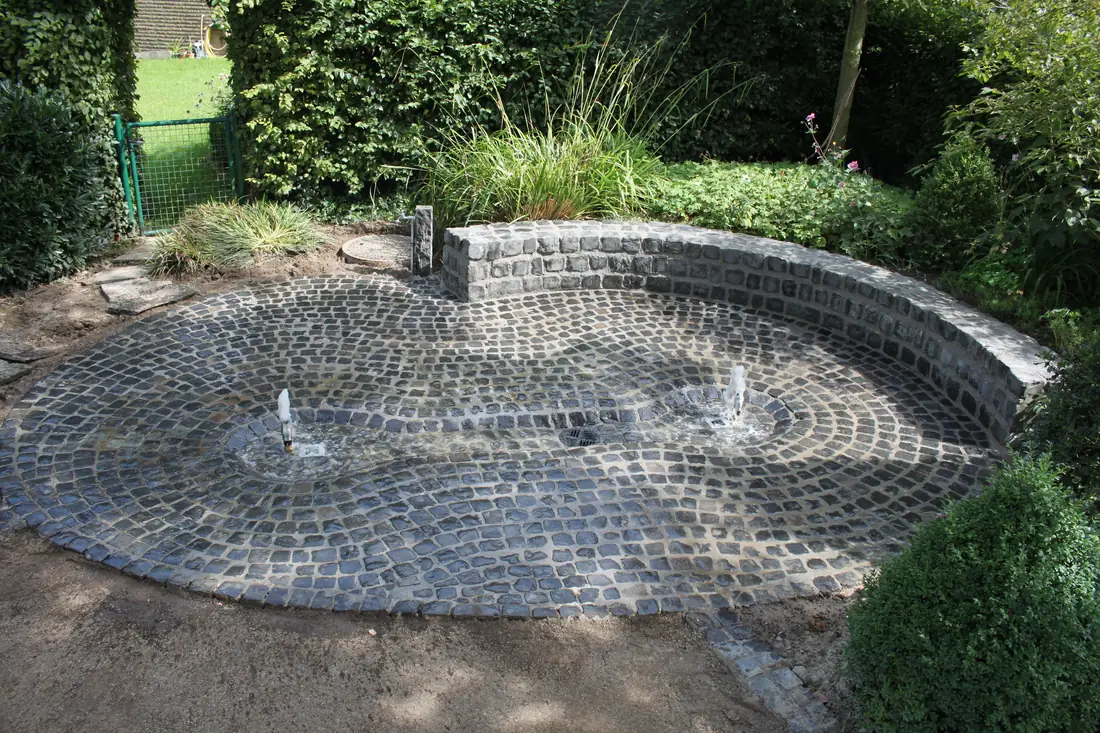 Rundes Wasserspiel aus Kleinpflaster mit Fontänen und halbrunder Natursteinmauer